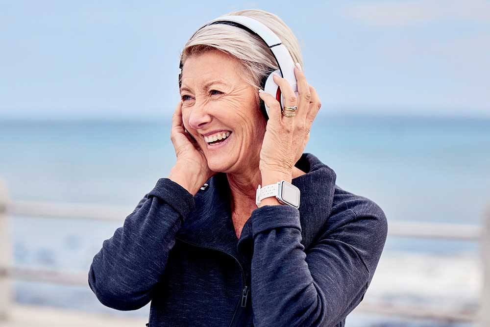 A senior woman standing on the beach listening to music on her headphones